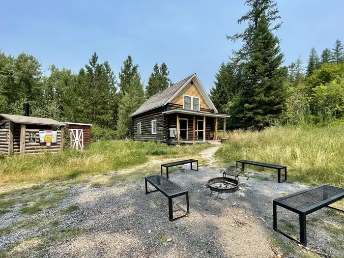 Swan Guard Station exterior. Fire ring and benches in the foreground. Cabin, vault toilet, and firewood shed in the background.