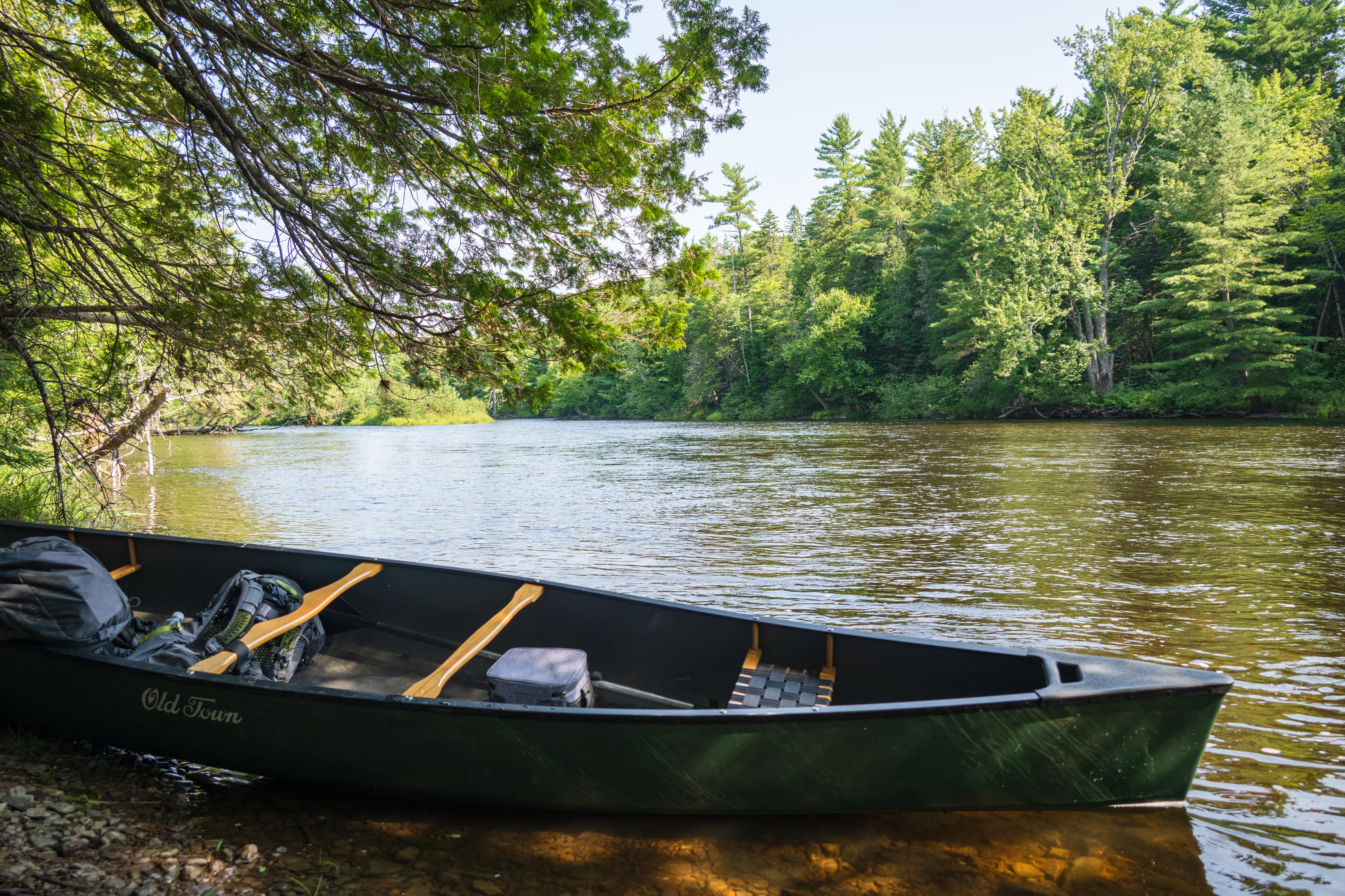 A green canoe with camping gear on the short gravel shore with the flowing river behind it.