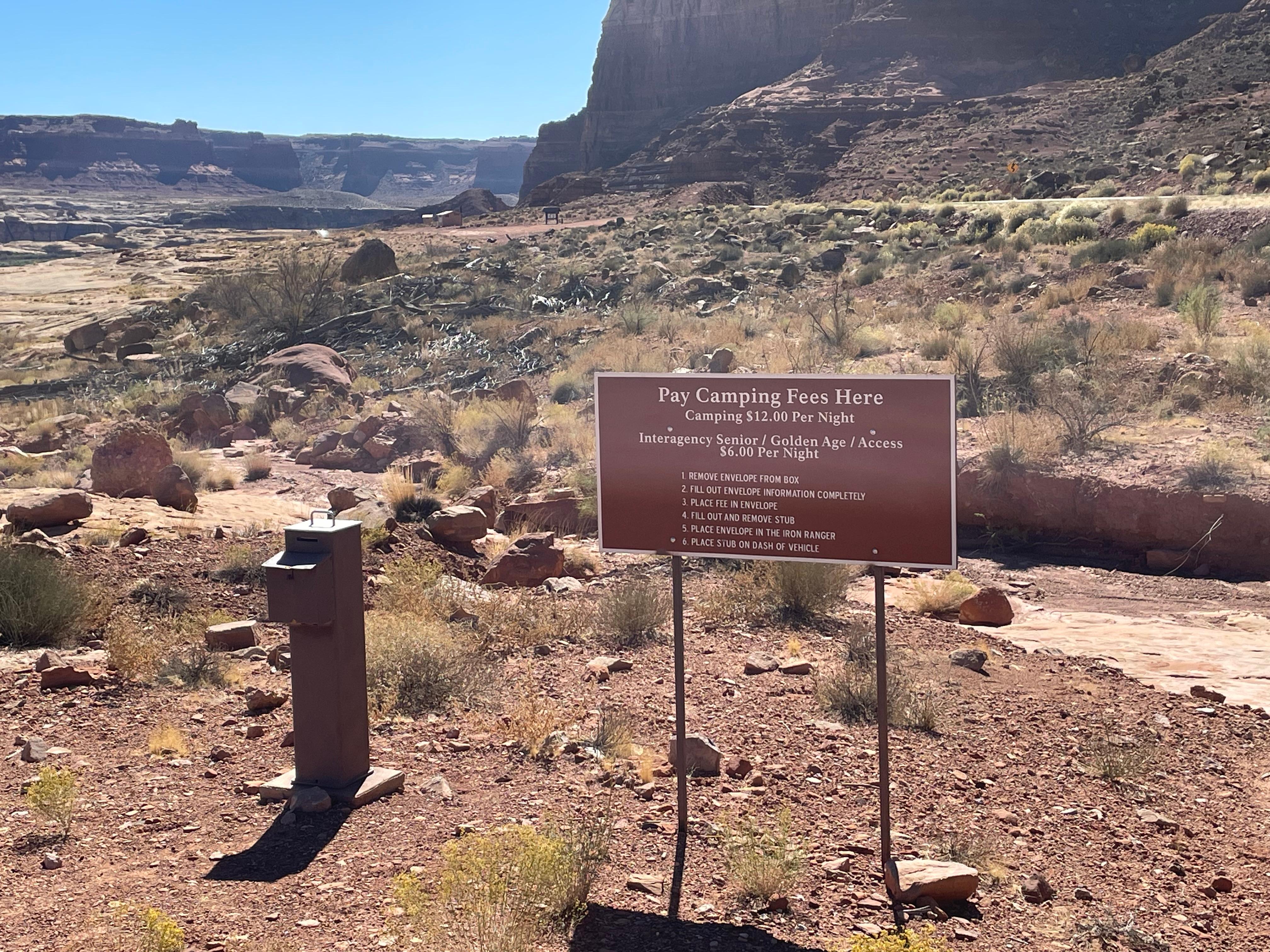 A rocky desert landscape. A stout metal post with a slot in the top is next to a sign: Pay camping F