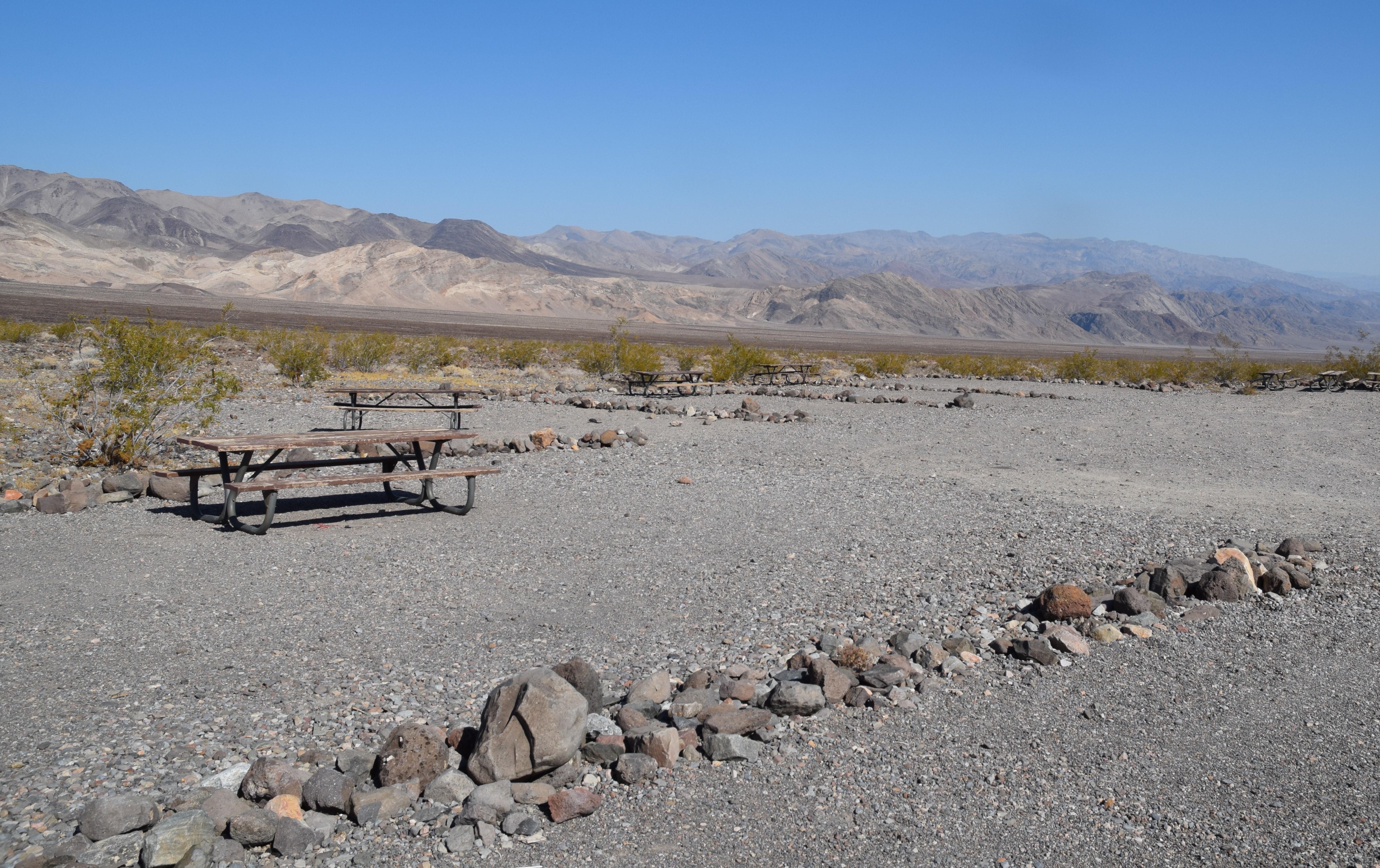 Row of loose 12 inch rocks separate a flat, gravel area. Metal picnic bench on far side.