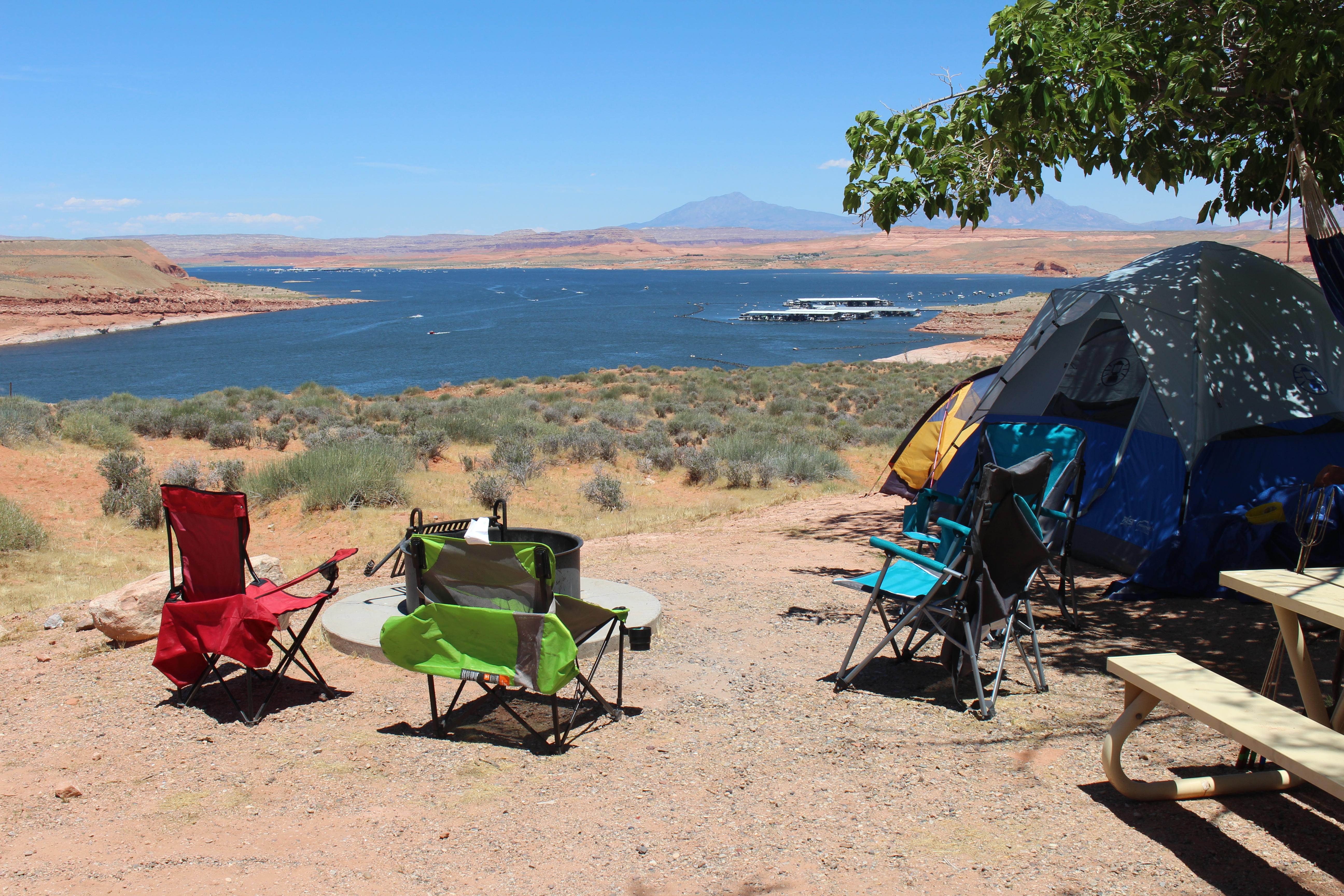 Three colorful lawn chairs and a rounded tent sit under a tree overlooking a desert lake